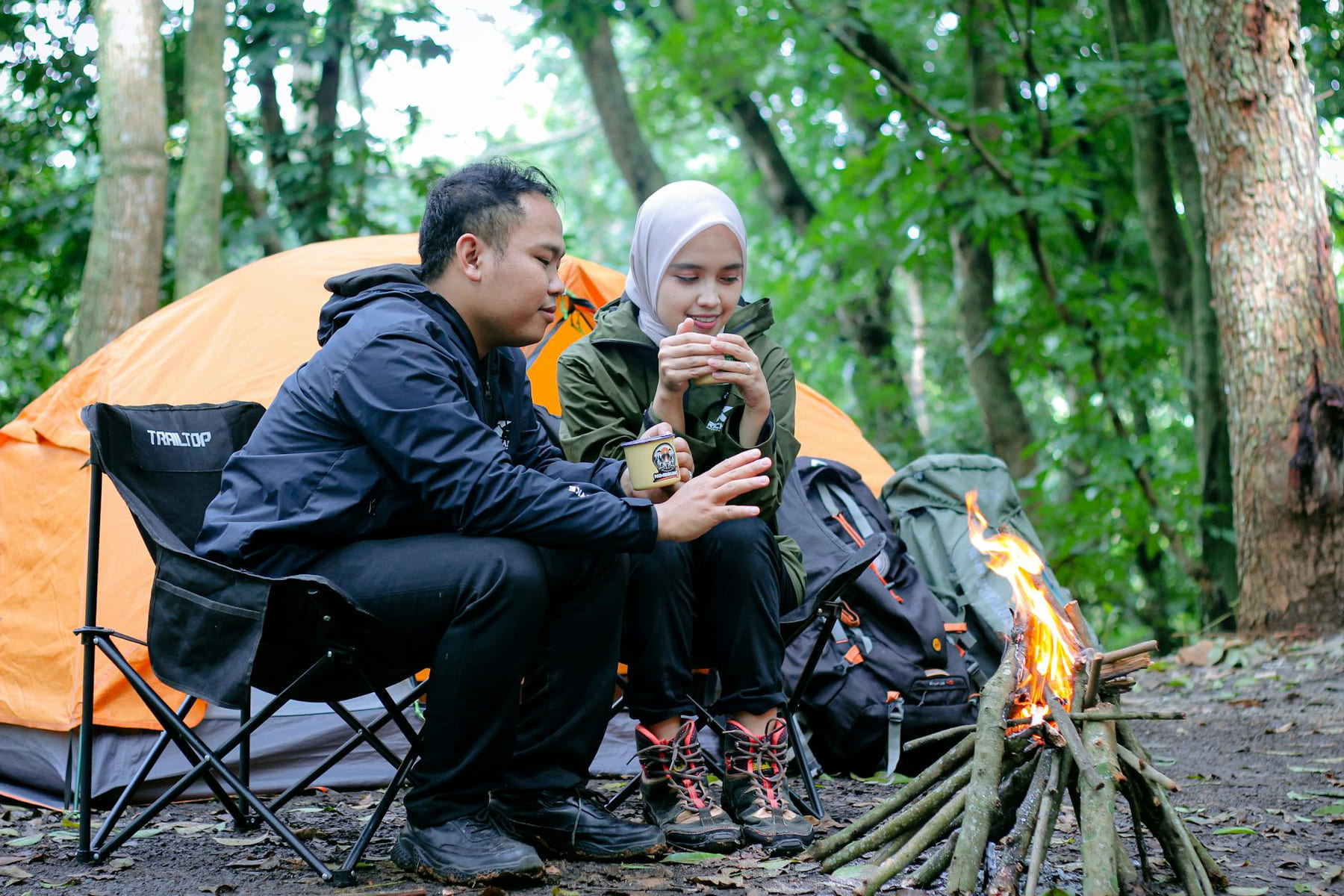 A couple enjoys a warm campfire in a forest campsite, camping near an orange tent. on The Frugal Camper