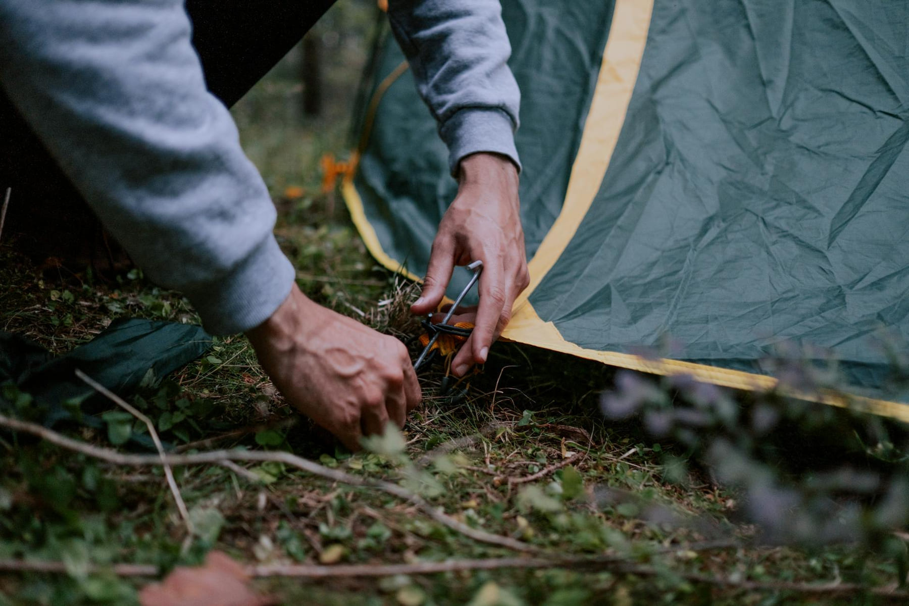 A close-up of a person securing a tent peg in a forested campsite, depicting outdoor camping activities. on The Frugal Camper