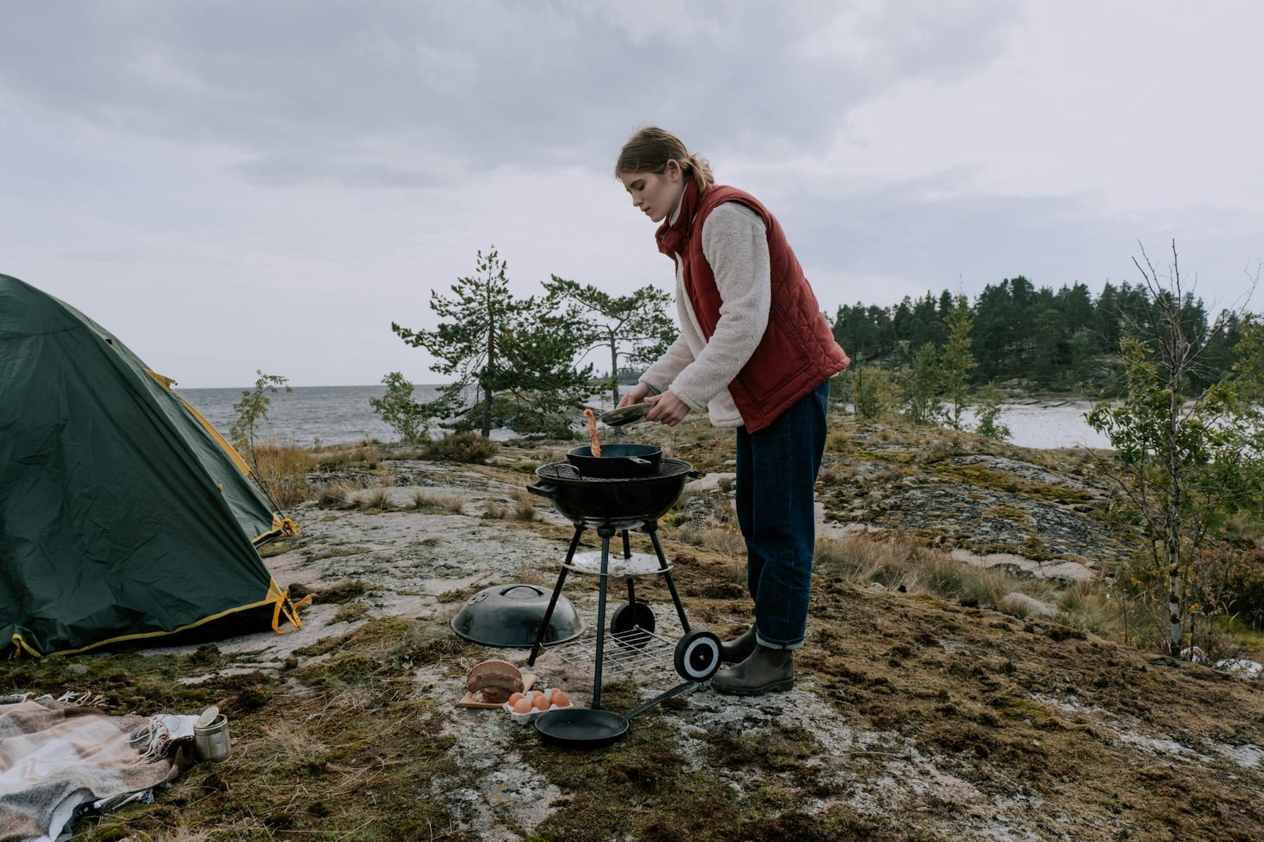 A woman cooks on a portable grill at a scenic seaside campsite. on The Frugal Camper