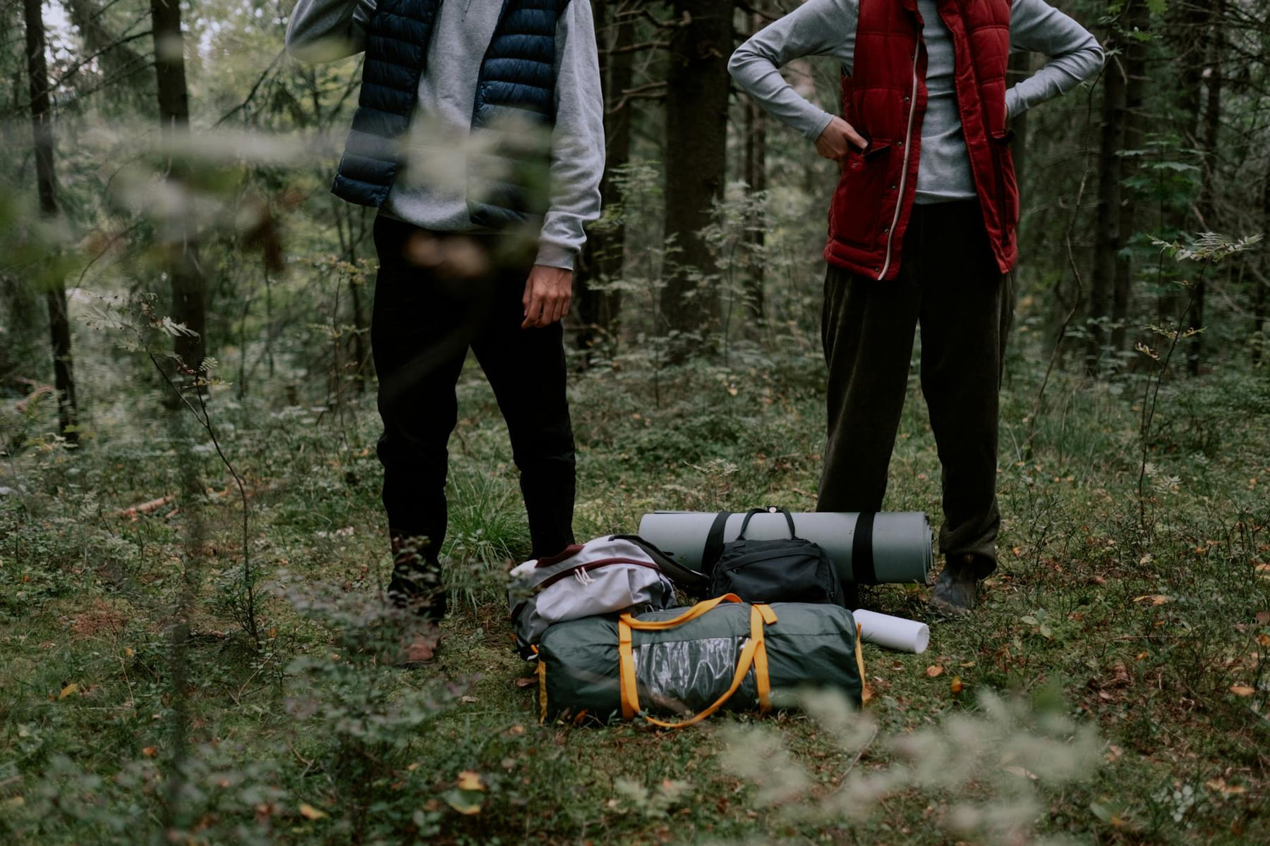 Two adults standing with camping gear in a forest, surrounded by greenery. on The Frugal Camper