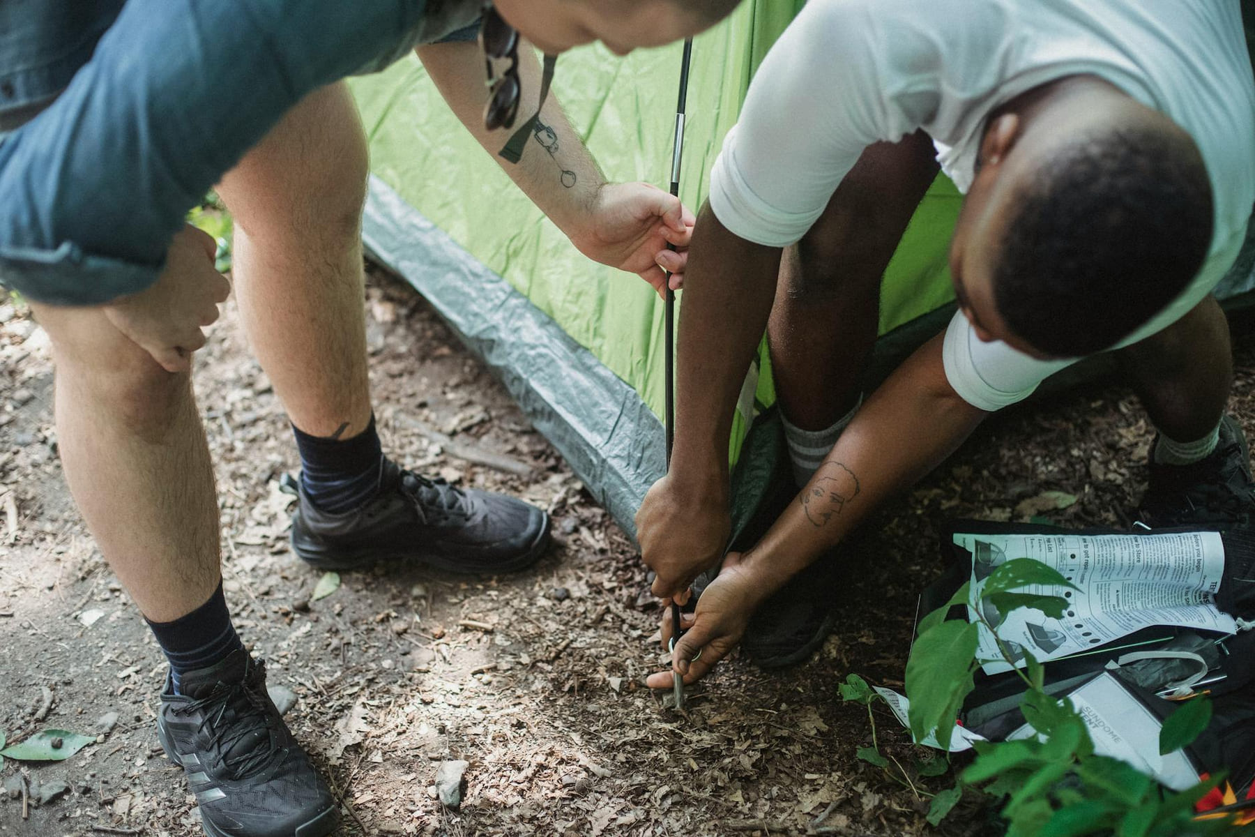 Unrecognizable black man setting up green tent with partner on ground in summer forest on The Frugal Camper