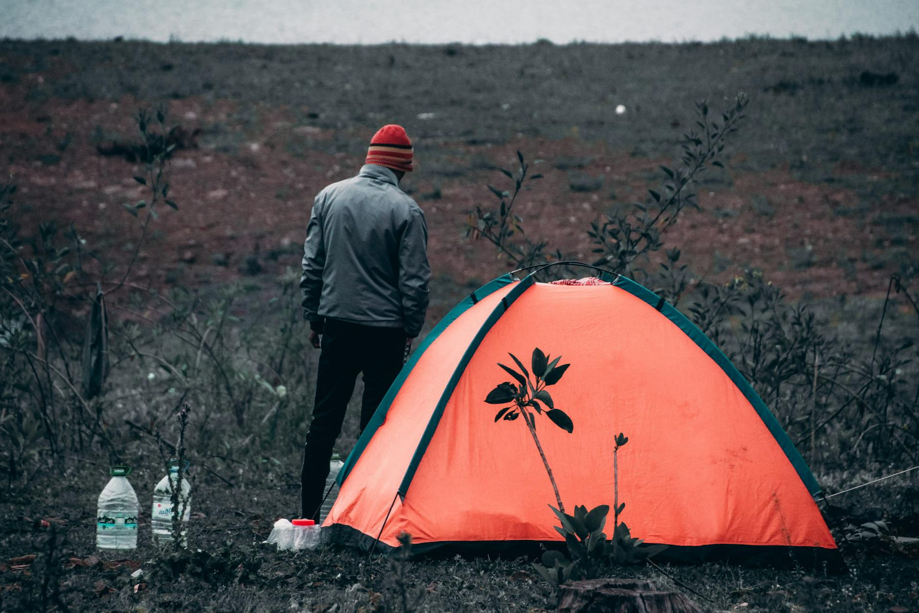Man in grey jacket camping in wilderness beside orange tent, enjoying outdoor adventure. on The Frugal Camper